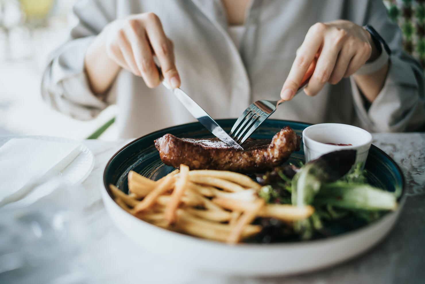 Close up of young woman cutting a juicy, grilled fillet steak on her plate on dining table, enjoying lunch in an outdoor restaurant. Outdoor dining. Lifestyle, people and food concept