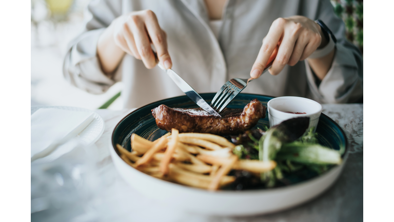 Close up of young woman cutting a juicy, grilled fillet steak on her plate on dining table, enjoying lunch in an outdoor restaurant. Outdoor dining. Lifestyle, people and food concept