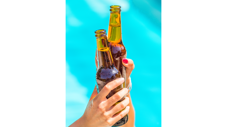 Female friends cheer clinking bottles of beer in their hands next to a pool of blue water.