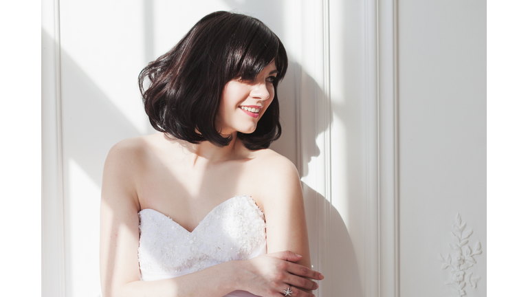 Portrait of young attractive brunette with short hair in a wedding dress. She is standing at the white walls, free space on the right. Joyful bride.