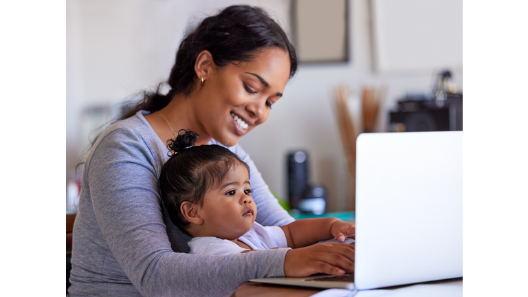 Working mother and baby at home, using a laptop. Young woman browsing online on a computer while her cute little girl is sitting on her lap. Happy mom and daughter on maternity leave or remote work