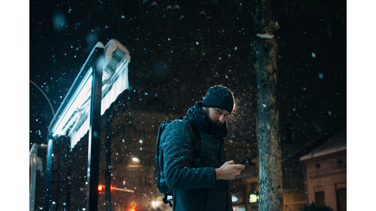 Man Using Mobile Phone While Standing Against Building During Snowfall At Night