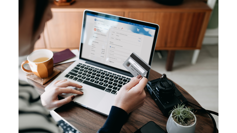Over the shoulder view of young Asian woman shopping online for flight tickets on airline website with laptop, entering credit card details to make mobile payment at home. Camera and passport on the table. Travel planning. Booking a holiday online