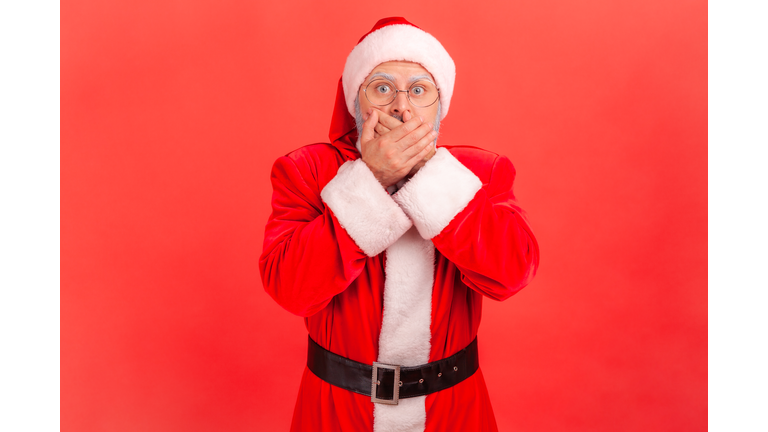 Portrait of elderly man with gray beard wearing santa claus costume closing mouth with his hands, afraid to say too much, embarrassment.