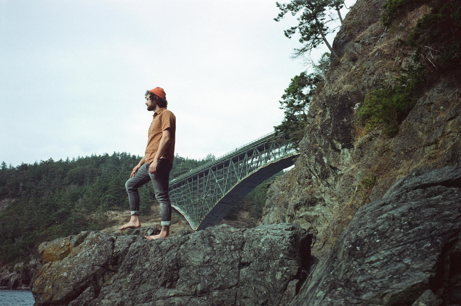 Low angle iconic portrait of a man standing on a rock formation with Deception Pass Bridge in the background