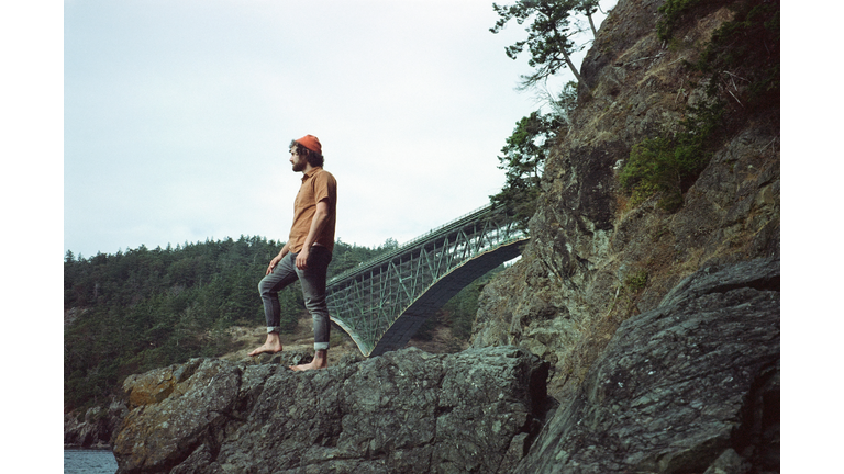 Low angle iconic portrait of a man standing on a rock formation with Deception Pass Bridge in the background
