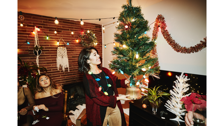 Smiling woman holding miniature Christmas tree during holiday party with friends