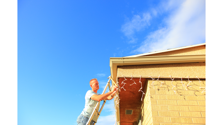 Man Hanging Holiday Lights On House With Blue Sky