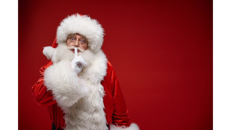 Kind Santa Claus shows us keep a secret sign with his finger on lips, standing on a bright red background with a copy space on a half of the photo