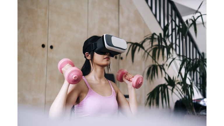 Young woman using virtual reality headset while exercising with dumbbells at home