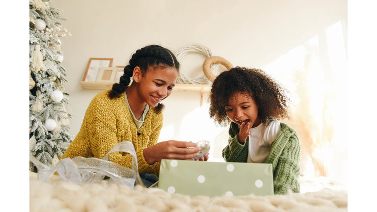 Two Afro sisters sit on a large bed and open a gift. Room design in light colors. Christmas mood. Christmas presents.