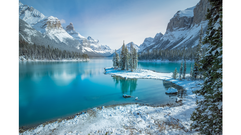 Snow covered land and trees surrounding a lake with a snow capped mountain in the background.