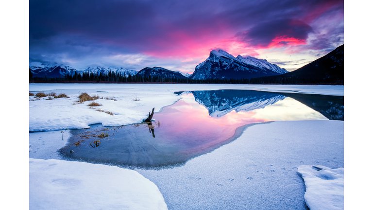 Pink sunrise over mountain reflected in partially frozen lake with snow