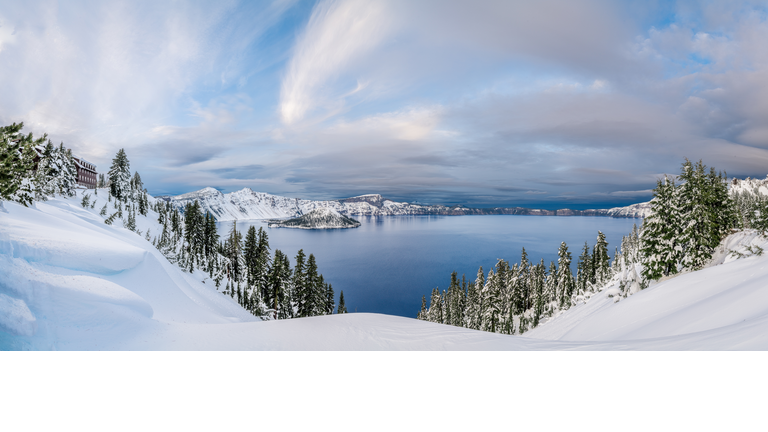 Fresh snow covers the areas surrounding Crater Lake, Wizard Island is seen on the left, and dramatic clouds cover the sky.