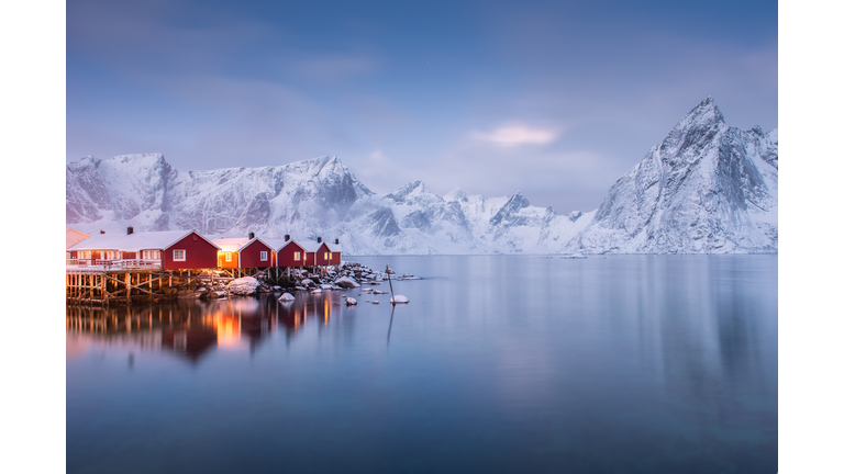 A small village juts out over a lake with snowy mountains in the background.