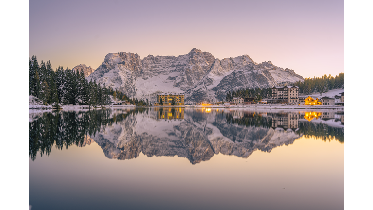 Sunrise tingeing the sky, snowy mountain, and lake in a pale pink.