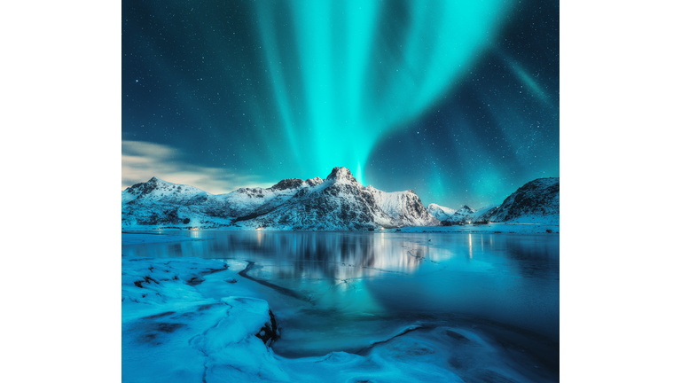 Aurora borealis over snowy mountains, frozen sea coast, reflection in water at night. Lofoten islands, Norway. Northern lights. Winter landscape with polar lights, ice in water. Starry sky with aurora