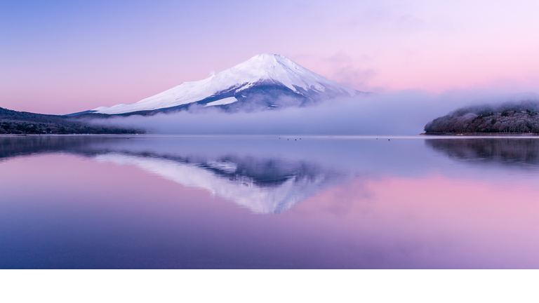 Dramatic snowy mountain peak rising above a placid lake and the pink sunrise colors the sky and water.
