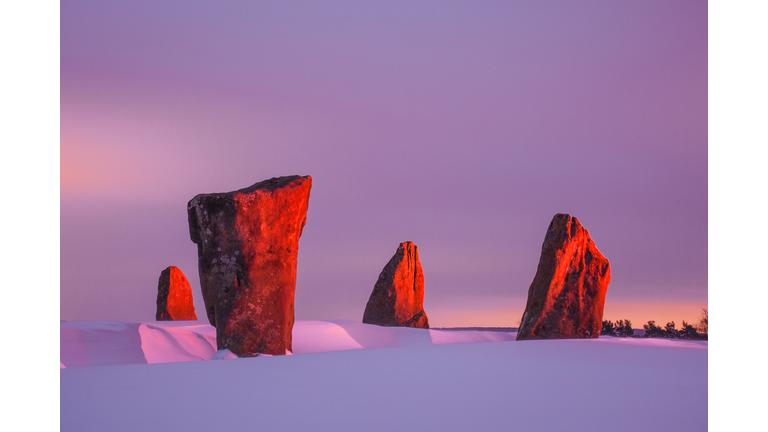 A red dawn sharply outlining the dark dramatic stones jutting out of the white snow.
