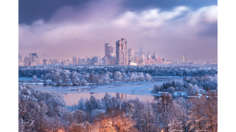 Aerial view of winter Moscow at evening dawn, snowed natural parkland, Moskva river and high-rise residential districts on the background, with picturesque weather conditions