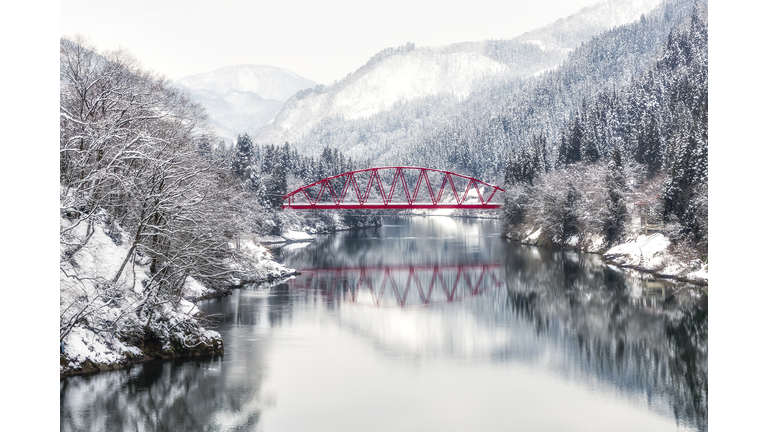 A red bridge dramatically stands out against the frozen river and snowy landscape with a mountain in the background.