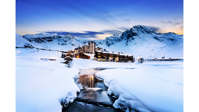 A river and a ski resort standing out starkly against the snow covered land in the forefront and the mountains behind it.