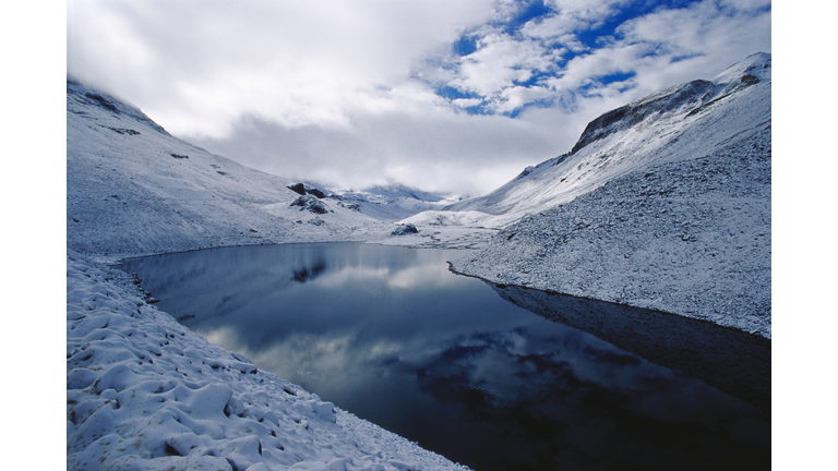 A dark river flowing through a snow-covered landscape starting from the right lower corner of the image and then curling back and disappearing around a snow covered mountain.
