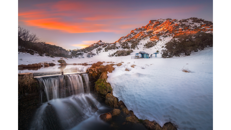 Waterfall and snowy mountain against a dramatic sunset sky.