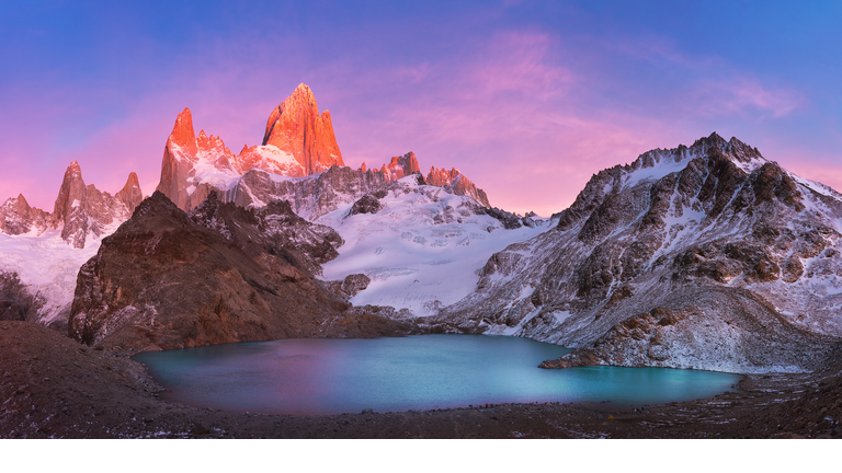 Dramatic red peaks jutting out of the snow above a placid lake.