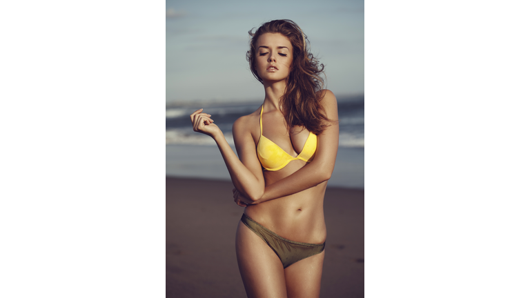 Brunette in a two-toned bikini, yellow top and brown bottom, posing with attitude on the beach.