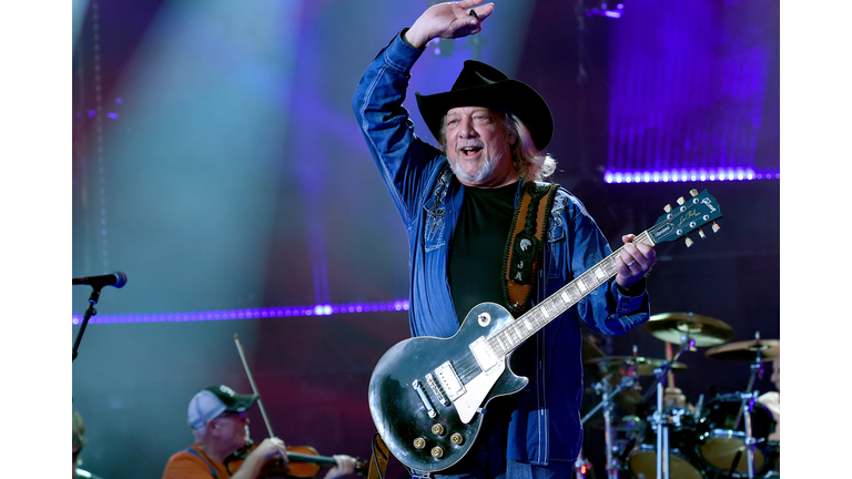 John Anderson in a jean jacket, black shirt, and black cowboy hat, with on arm raised over his head and a guitar.