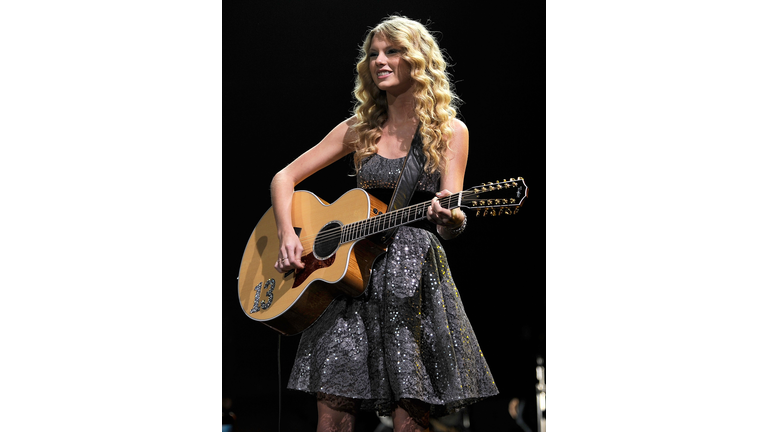 Taylor Swift with long curly blonde hair, wearing a gray sparkly dress, and holding a guitar.