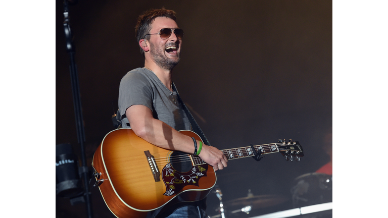 Eric Church wearing a gray t-shirt and sunglasses while laughing and holding a guitar.
