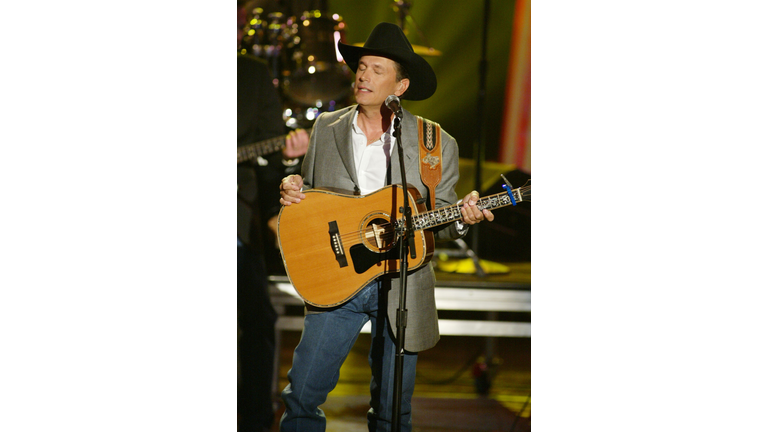 George Strait, wearing jeans, a khaki jacket, and a black cowboy hat, singing into a microphone while holding a guitar.