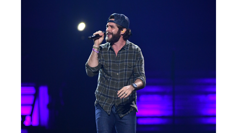 Thomas Rhett sporting a beard, plaid shirt, backwards ball cap, and jeans, singing into a microphone.