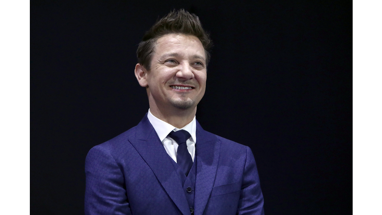 A smiling Jeremy Renner in a dark blue suit against a black background.