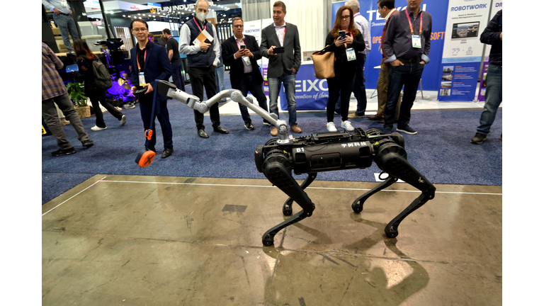 Black four-legged robotic dog displayed on a wooden space with an audience in the background.