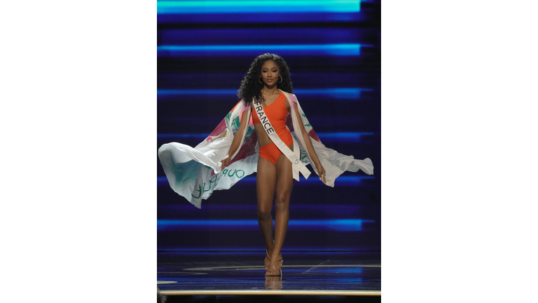 Curly-haired, dark-skinned brunette in orange swimsuit and white cape.