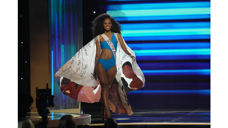 Dark-skinned brunette in blue swimsuit and cape that is brown on one side and white on the other.