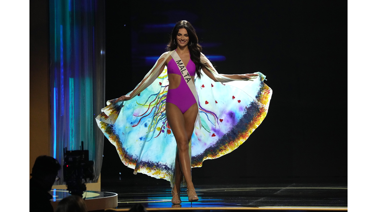Brunette in a purple swimsuit and multi-colored cape.