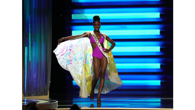 Dark skinned brunette in lavender swimsuit and lavender, yellow, and blue cape.