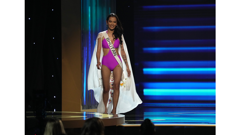 Dark-skinned brunette in purple swimsuit and white cape.