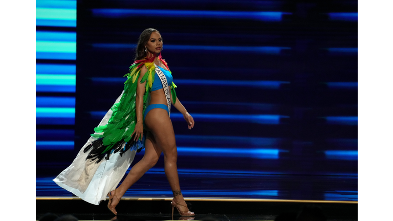 Brunette in blue swimsuit and white cape with green and black feathers.