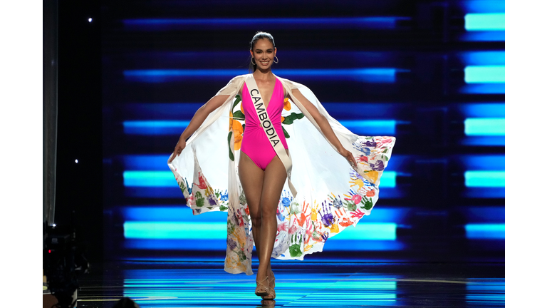 Brunette in pink one-piece swimsuit and white cape with multi-colored handprints along the border.