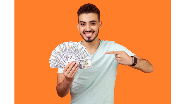 Portrait of lucky rich brunette man pointing at dollar banknotes. indoor studio shot isolated on orange background