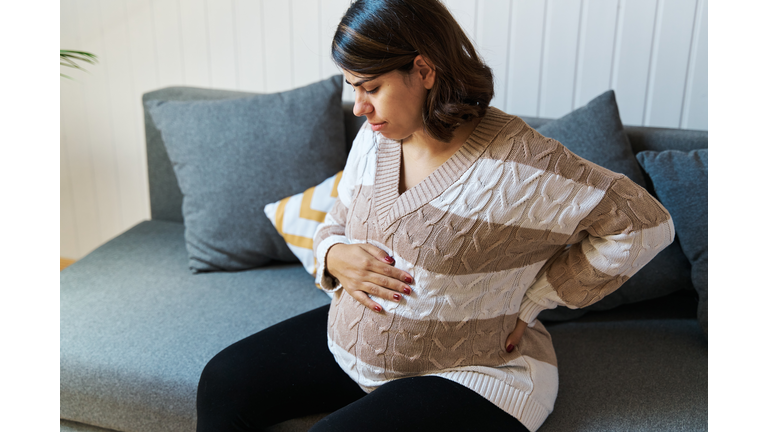Pregnant woman feeling tired and suffering back pain while sitting on a couch at home.
