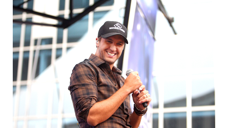 Luke Bryan in a plaid brown shirt and baseball cap holding a microphone and smiling.