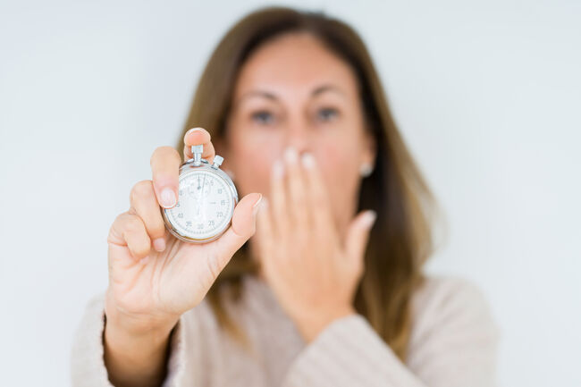 Middle age woman holding stopwatch isolated background cover mouth with hand shocked with shame for mistake, expression of fear, scared in silence, secret concept