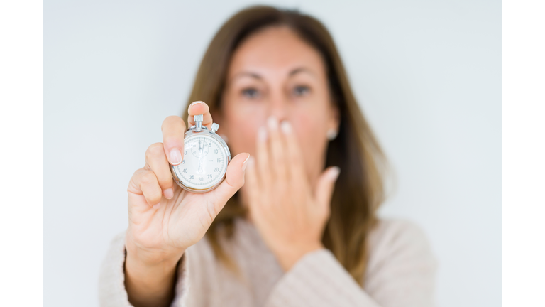 Middle age woman holding stopwatch isolated background cover mouth with hand shocked with shame for mistake, expression of fear, scared in silence, secret concept