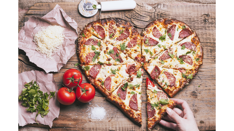 Hand pulling slice of heart-shaped pizza near ingredients on cutting board